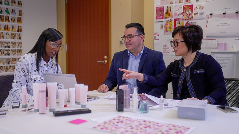 Three people collaborating at a table with skincare products, laptop, and design mockups