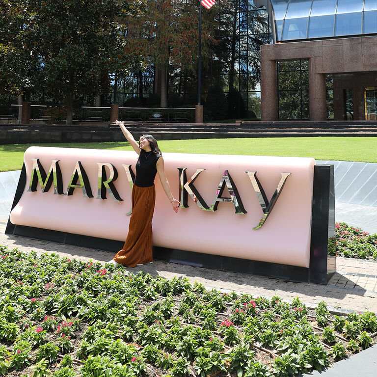 Person standing beside a large Mary Kay sign outdoors with landscaped flowers and office building in background